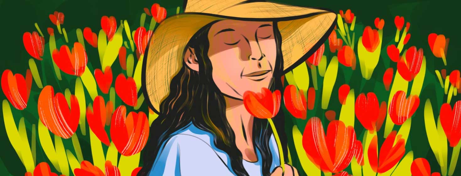 A woman in a sunhat closes her eyes and smells a flower she is holding, while standing in a field of red flowers.